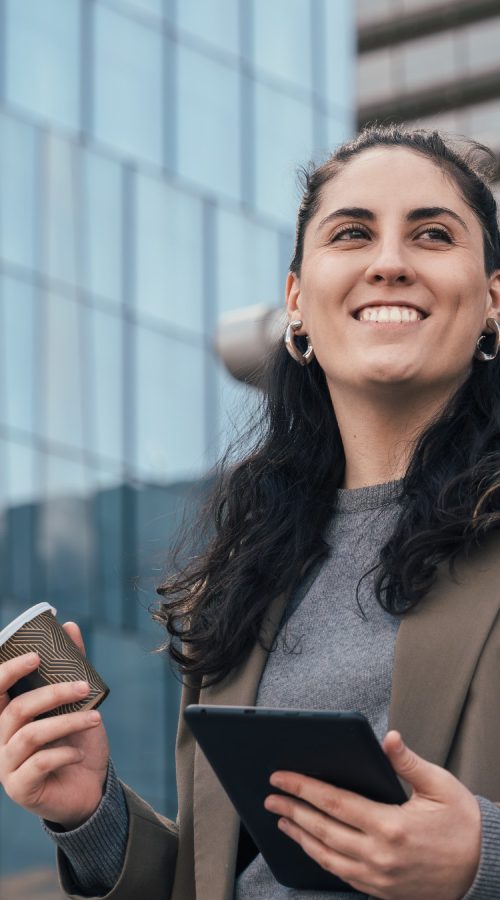 A woman in professional attire standing outside of an office building holding a tablet while smiling