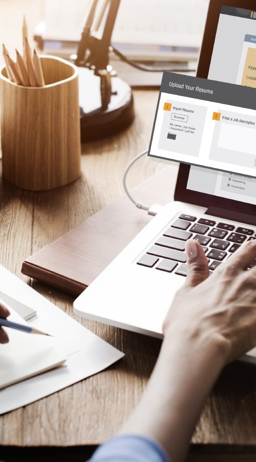 A woman sitting at a desk typing on a laptop with a notebook next to her. On the laptop screen it has an icon that says 'job search'