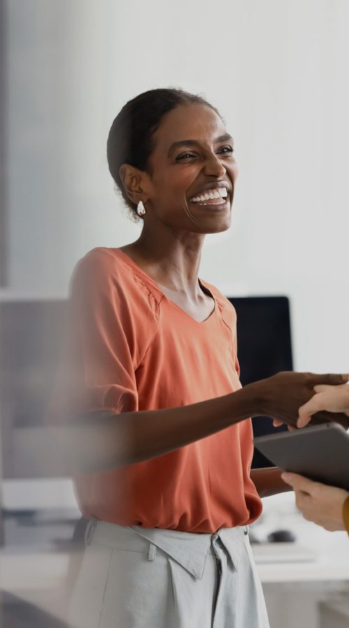 two women in business attire shaking hands while smiling. Background is an office setting.