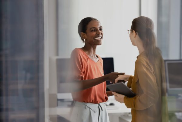 two women in business attire shaking hands while smiling. Background is an office setting.