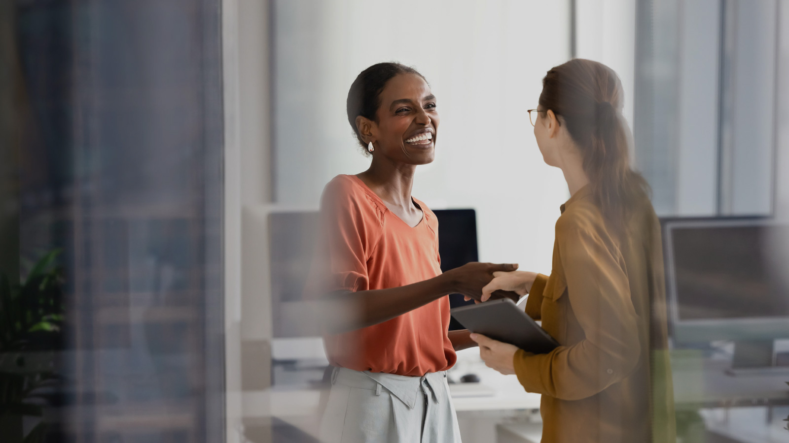 two women in business attire shaking hands while smiling. Background is an office setting.
